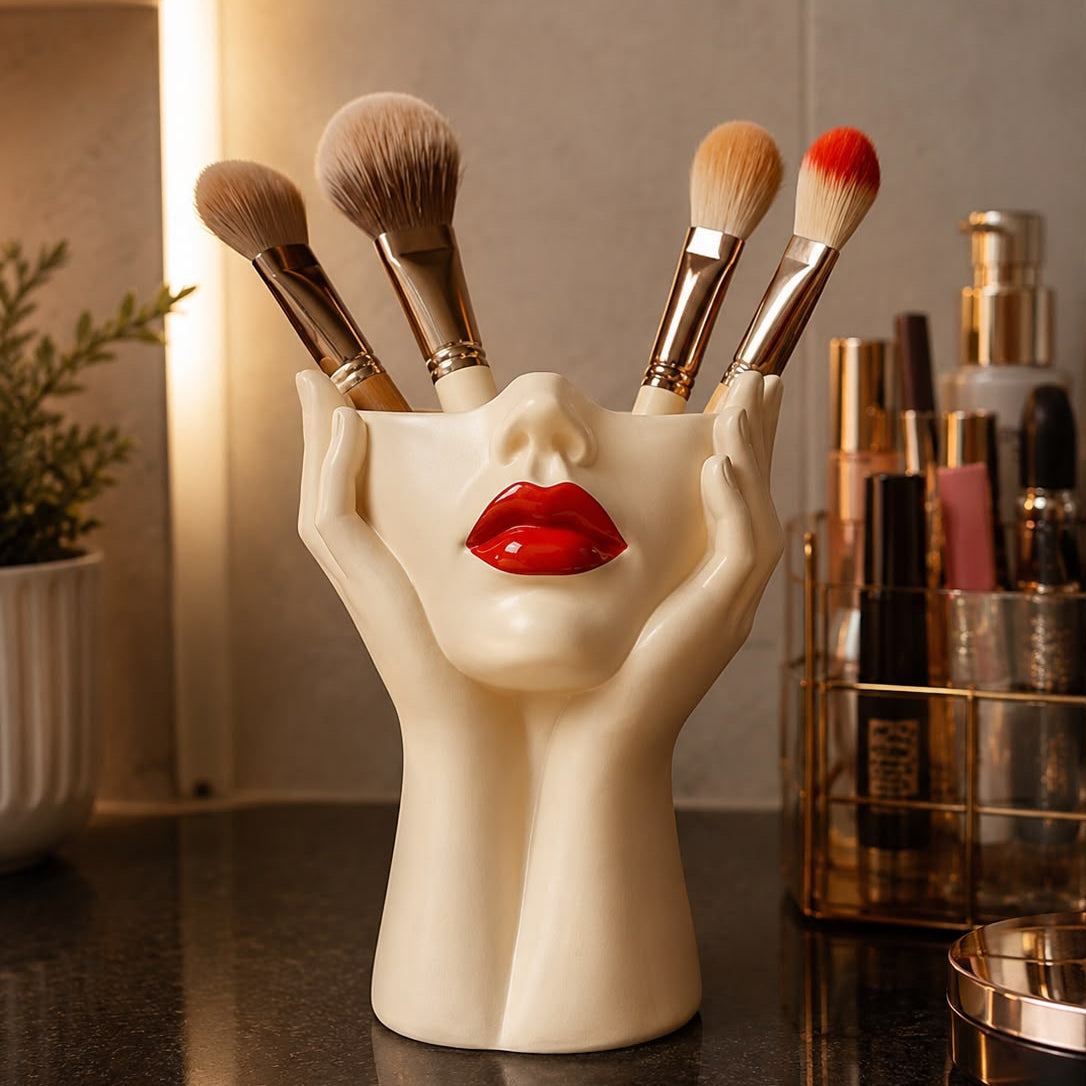 A sculptural makeup brush holder shaped like a woman's face and hands, holding several makeup brushes, set on a dark countertop with cosmetic bottles in the background.