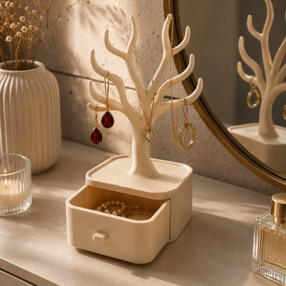 A cream-colored tree-shaped jewelry organizer with a small drawer base, styled on a vanity table with earrings hanging from its branches and a round mirror behind it.