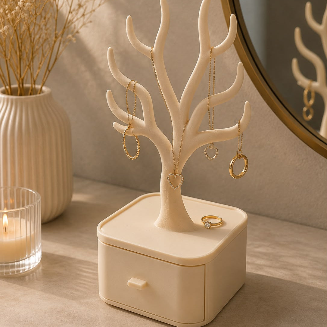 A cream-colored jewelry tree display stand with a small drawer base, styled on a tabletop with gold hoop and chain earrings hanging from its branches, next to a candle and dried flowers.