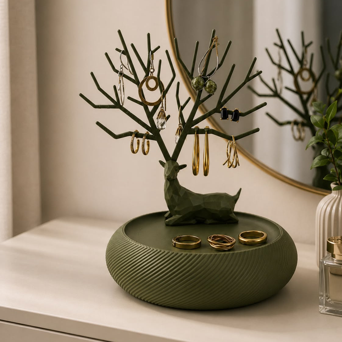 An olive green jewelry organizer with a deer-shaped tree stand and a round textured base, styled on a white surface with gold rings and earrings in front of a mirror.