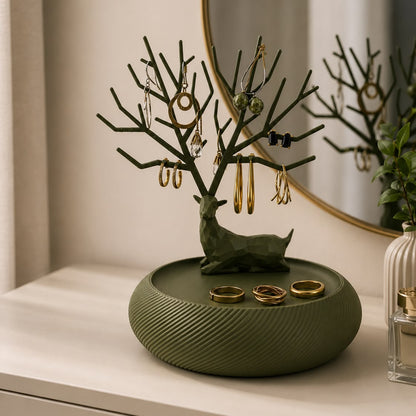 An olive green jewelry organizer with a deer-shaped tree stand and a round textured base, styled on a white surface with gold rings and earrings in front of a mirror.