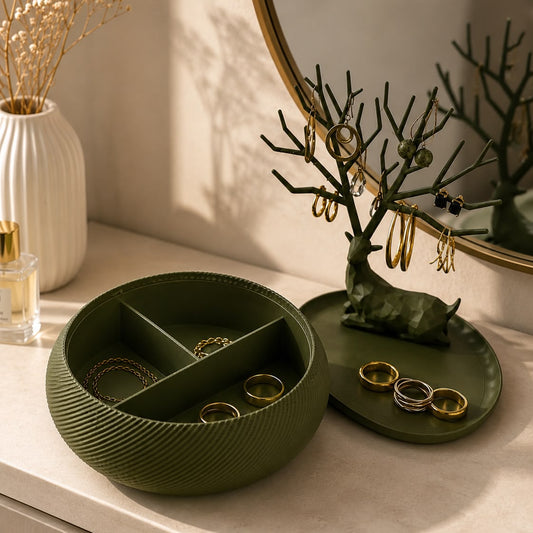 An olive green jewelry organizer set on a light marble vanity surface, featuring a round segmented bowl and a matching deer-shaped branch stand holding gold and silver jewelry, with a mirror and dried flowers in the background.