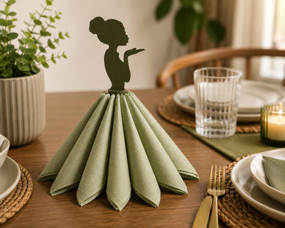A folded sage green napkin arranged in a skirt-like shape on a wooden dining table, topped with a dark silhouette cutout of a woman's profile. The setting includes plates, a glass, a candle, and a potted plant in a softly lit indoor environment.